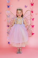 Smiling girl in fairy costume and tiara stands with wings on pink