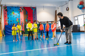 Male coach showing floorball technique to elementary school students wearing colorful vests in a...