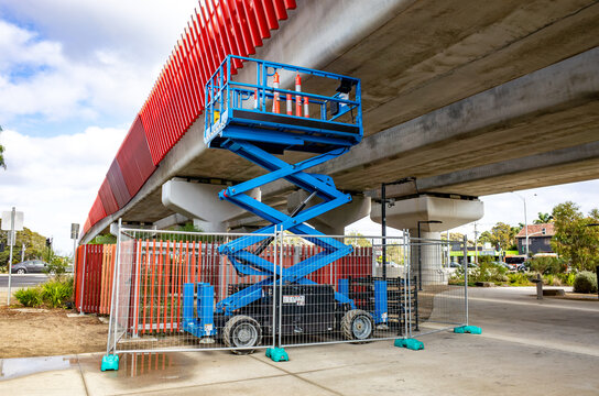 A scissor lift or elevated platform positioned underneath a concrete bridge or overpass, with maintenance work being conducted. - Powered by Adobe
