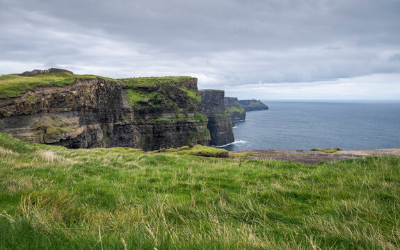 The Cliffs of Moher on Ireland&rsquo;s west coast, with green grass leading to the cliff edge under a grey sky, showcasing the natural landscape and geological features