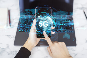 Person using smartphone with 5G technology hologram over laptop on light desk, representing global internet and data network connection concept.