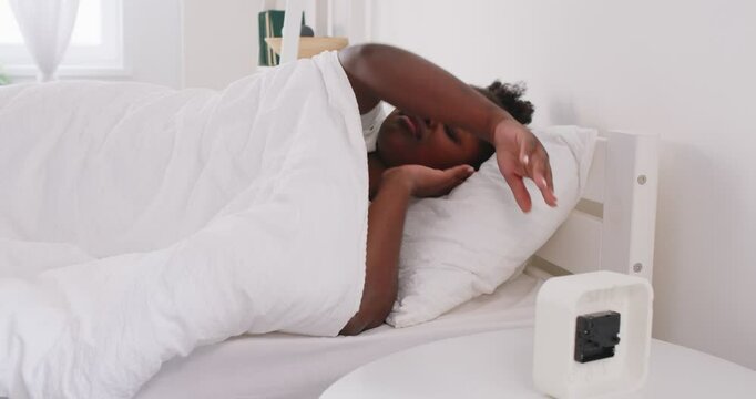 Sleepy young african american woman lying in bed in her cozy modern bedroom under white blanket, looking at alarm clock cant waking up early in the morning. Female person waking up. 4k video.