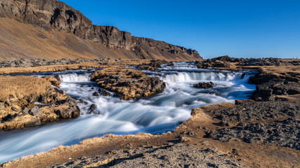 Langzeitbelichtung des Fossalar im Winter, blaue weißer Fluss durchzieht karge Berglandschaft, sonnig bei blauem Himmel