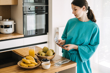 Young woman counting US dollars beside a kitchen counter with fruit and juice. Budgeting in the wellness era, conscious spending, financial self-care, food cost planning