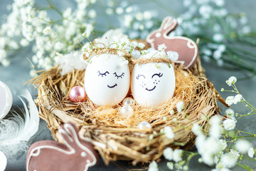 Two chicken eggs with hand drawn happy faces with wreaths in a nest of dry grass with feathers, rabbits and gypsophila branches on a light blue background.