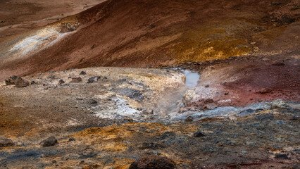 farbenfrohe Detailaufnahme im Geothermalgebiet Krysuvik-Seltun in Island, Dampf steigt auf, die umgebenden Hügel zeigen verschiedene Rot- und Brauntöne