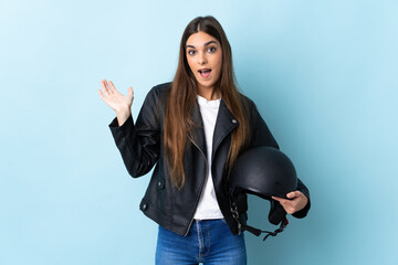 Young caucasian woman holding a motorcycle helmet isolated on blue background with shocked facial...