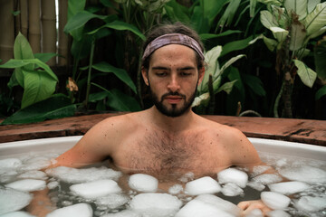 Man takes an ice bath outdoors in a lush tropical setting. Cold immersion for mental strength,...