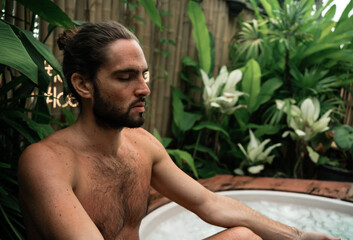 Man takes an ice bath outdoors in a lush tropical setting. Cold immersion for mental strength,...