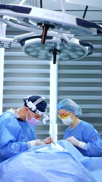 Medics leaned over the patient standing on two sides. Big round lamps on the ceiling above the doctor and nurse. Vertical video.