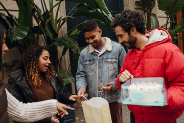 Two adult Latino men and women, stand surrounded by greenery in front of a white building with presents. The men wear a denim jacket and a red puffer jacket, while the woman wears a brown sweater.