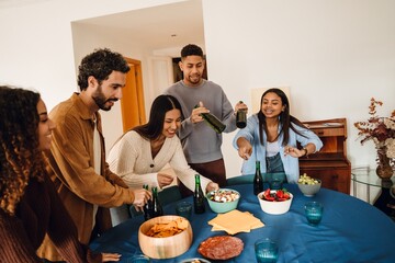 A young Black woman in her late 20s, instructing a diverse group of her friends while they set a small table with drinks and snacks, as they stand together in a cozy home