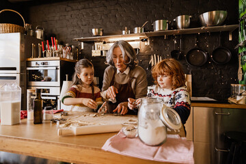 An elderly White grandma in her 70s, cutting shapes from dough together with her two school-aged...