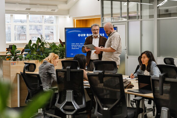 Fototapeta premium A Caucasian man in his 30s standing with an elderly Caucasian man with a beard in eyeglasses and looking at a clipboard, a multiethnic group of female adult colleagues sitting at a table in the office