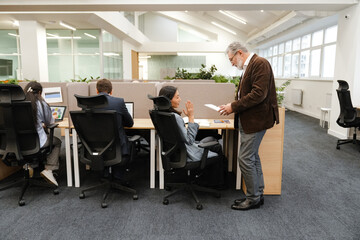 In an office, a gray-haired White man in his 60s stands and shows documents to his Asian female colleague in her 30s sitting at her desk and talking, with other workers at their desks near them.
