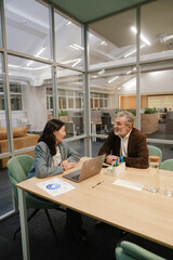 A senior White man with gray hair and a beard, wearing a brown jacket, talks with an Asian female colleague in her 30s at a wooden table with a laptop in a modern office meeting room with glass walls.