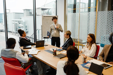 A Caucasian male intern in his late 20s, giving a presentation to a table of his older colleagues as he stands inside of an office meeting room, with a clipboard in hand and a whiteboard behind him