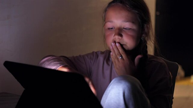 A girl sits at a table, staring at a tablet screen with a tired, bored expression. She yawns while looking at the screen. Great for themes of fatigue and technology use.
