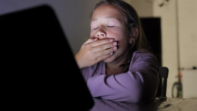 A young girl sits at a table, looking at a tablet screen. She appears tired and sleepy, yawning as she struggles to stay awake. Perfect for themes of screen time and fatigue.