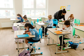 Multiethnic elementary school pupils are sitting at desks, drawing and writing, during a lesson, while their formally dressed Black female teacher in her 30s helps one of them, in a sunlit classroom.
