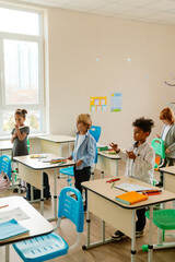 Obraz premium A young, school-aged White boy with short blond hair, who is dressed in a blue button-up shirt, standing next to his desk in a first-grade classroom, as his classmates do the same around him,