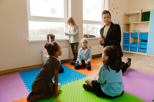 An adult Latino female teacher leads a learning activity with a diverse group of elementary school students in a classroom. The room includes colorful floor mats, shelves, and storage containers.
