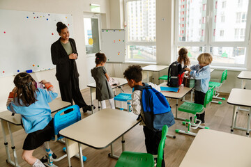 Diverse elementary school students and an adult Latino female teacher stand in a classroom. The students are standing up in the classroom with whiteboards, white windows, and wooden floors.