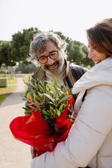 An elderly White man with a beard and glasses gifts a plant in a red package to an adult White...