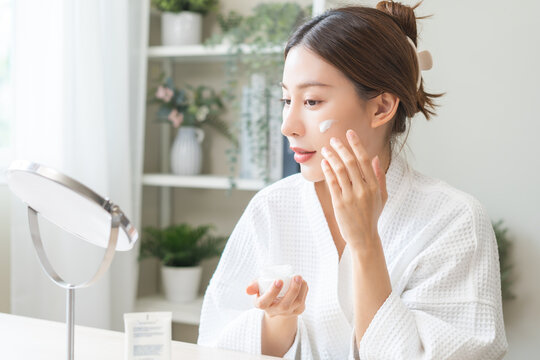 Facial beauty skin care, smile of pretty asian young woman in bathrobe looking at mirror, hand applying moisturizer lotion on her face, putting cream treatment before makeup cosmetic routine at home.