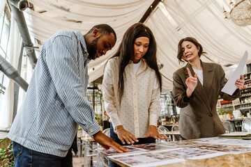 A cute Black couple in their late 20s, looking at the pictures of designs as they talk to their mid-30s White female wedding planner, while standing next to a marble table in a restaurant chamber