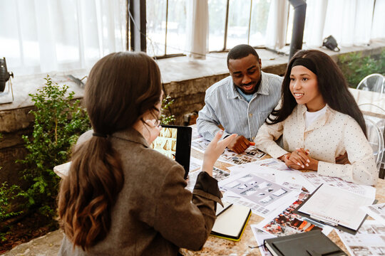 Two diverse women, and an adult Black man with short dark hair, sit at a table discussing photos and documents. Large windows and greenery are in the background. The people discuss a wedding.