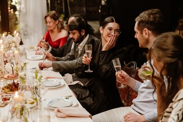 A multiethnic group of formally dressed adult people in their 30s is sitting at a long decorated table, talking, holding champagne glasses, smiling and laughing, celebrating a wedding in a restaurant.