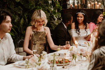 A young Black bride in her late 20s, dressed in her wedding dress, talking to her newlywed Black husband, as they sit at the dinner table among the guests, after their wedding