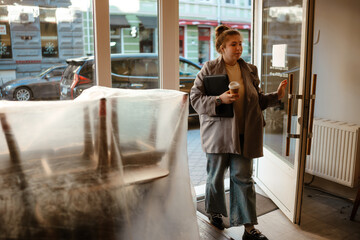 A young White woman with red hair in a bun wearing a gray coat holds a coffee cup and a tablet while walking into a store. A car is parked outside along with other storefronts.