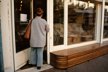 A view from the back of a young, overweight white-skinned woman dressed in a gray business suit, walking into a newly-built coffee shop from the street, with a purse over her shoulder
