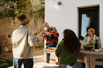 A White woman in her early 30s, with a bald head, laughing cheerfully as she talks to a diverse group of her adult friends as they all hang out on a small patio of a residential house, on a sunny day