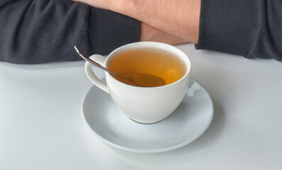 morning tea routine, a saucer holds a white teacup with tea and a spoon in the background, crossed arms may belong to someone in a dark sweater