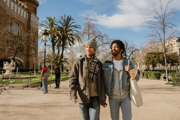 Obraz premium A cute gay couple of a Black man and a White man in their 30s, taking a stroll along a park together, looking around and bathing in the sunlight while holding hands, as they're on their date