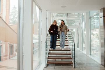 A young White woman in her late 20s, walking down a set of stairs with glass walls around them together with her young White female friend, talking to her and looking at her phone