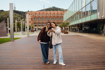 Two young White women with long hair stand in front of a historic ornate building beside a modern glass structure. They wear sweaters and jeans, and one holds a smartphone to take a selfie.
