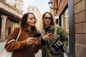 A adult woman with long blonde hair in eyeglasses and a jacket standing with a White woman in her late 20s with brown hair on a street with old building while they both looking at their smartphones