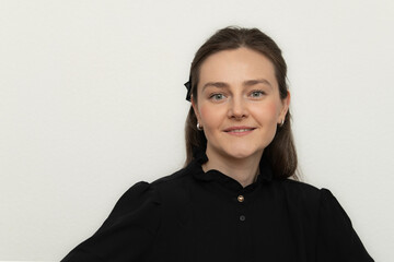 A woman with long hair and a black blouse stands in a studio, smiling warmly. The minimalistic background emphasizes her confident demeanor and professional appearance
