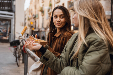 Two young White women stand on a city street. One wears a brown coat while the other wears a green jacket. The woman in the brown coat eats food from a brown paper plate as they converse.