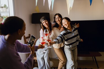 A Black woman in her late 20s in a purple sweater taking a photo of three multiethnic young adult women posing with glasses of cocktails in a living room with white walls and a TV during a home party