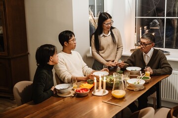 An Asian adult couple with black hair and a teenage son in a black sweater gathered at a table with an elderly Asian grandfather with short gray hair during a dinner with various meals in a room