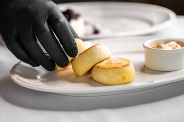 A chef in black gloves carefully places fluffy pancakes on a white plate, accompanied by a small dish of creamy sauce. The meticulous arrangement highlights the gourmet dessert.