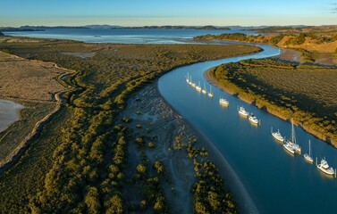 Aerial view of boats moored in a river, surrounded by lush greenery and mudflats, leading to the...