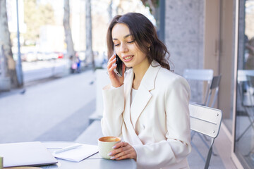 Beautiful young lady in elegant white jacket talking on the phone at a cafe in the city