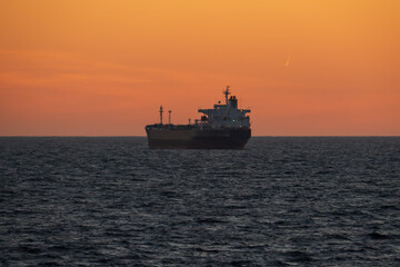 A large cargo ship sails on the dark sea against a beautiful orange sunset sky.