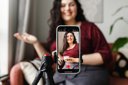 Selective focus on smartphone mounted on tripod recording female lifestyle blogger or influencer holding cup of coffee or tea gesticulating, giving advice on reducing stress and anxiety